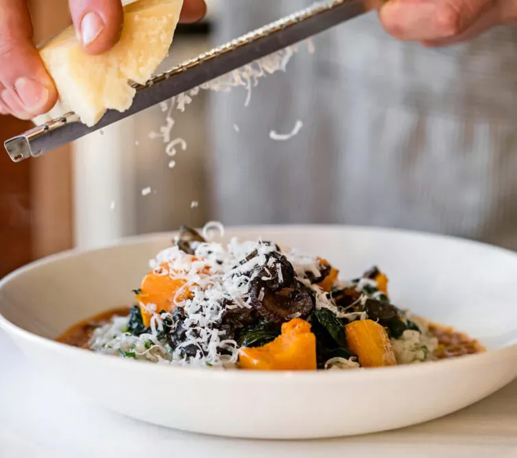 At Avalon Hotel, a person grates cheese over a dish of roasted vegetables and greens, beautifully served on a white plate.